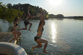 Two girls and a guy jumping off a dock into the lake.